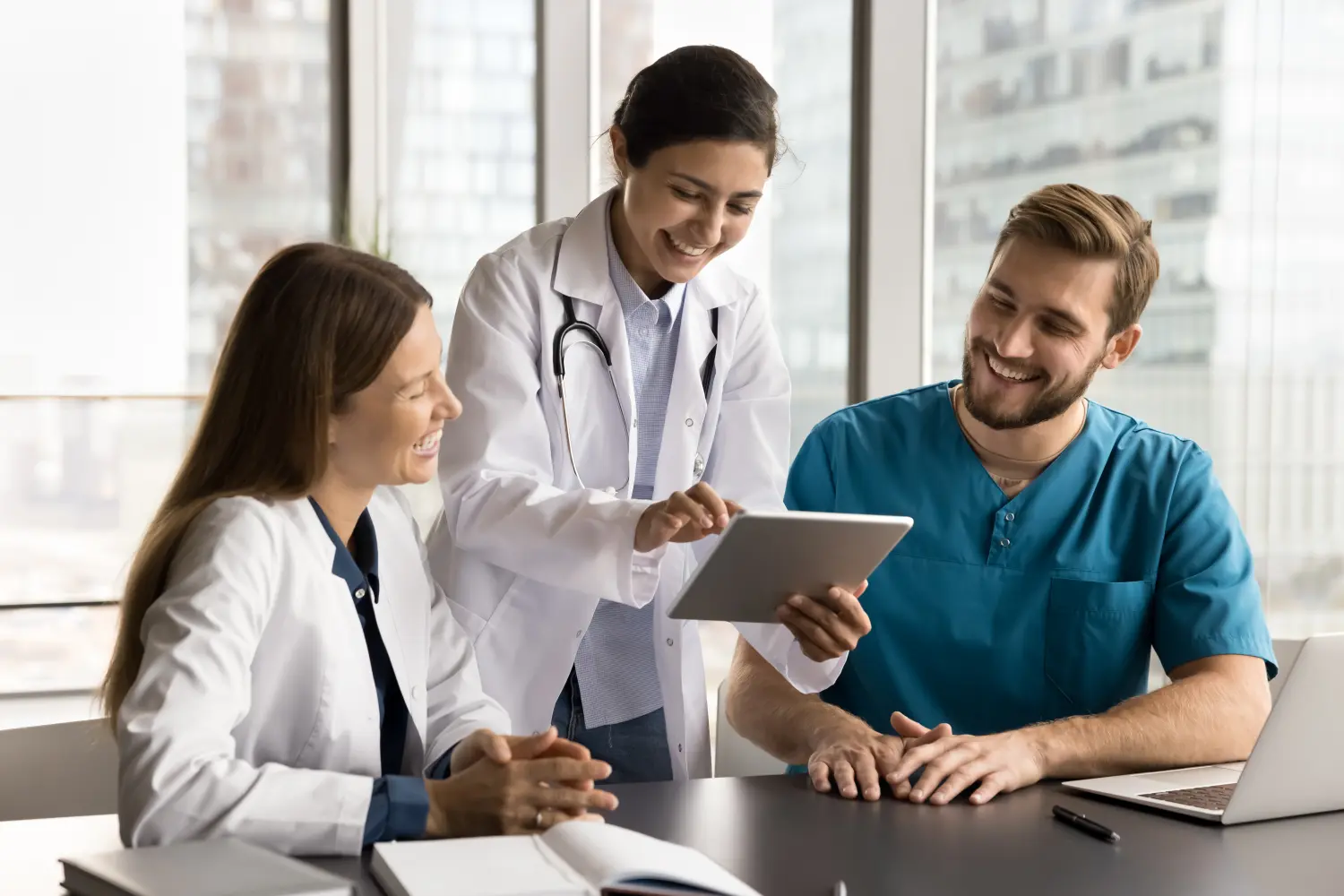 Fröhliche, vielfältige medizinische Kollegen in Uniformen aus Kliniken benutzen gemeinsam Tablet-Computer am Arbeitsplatz