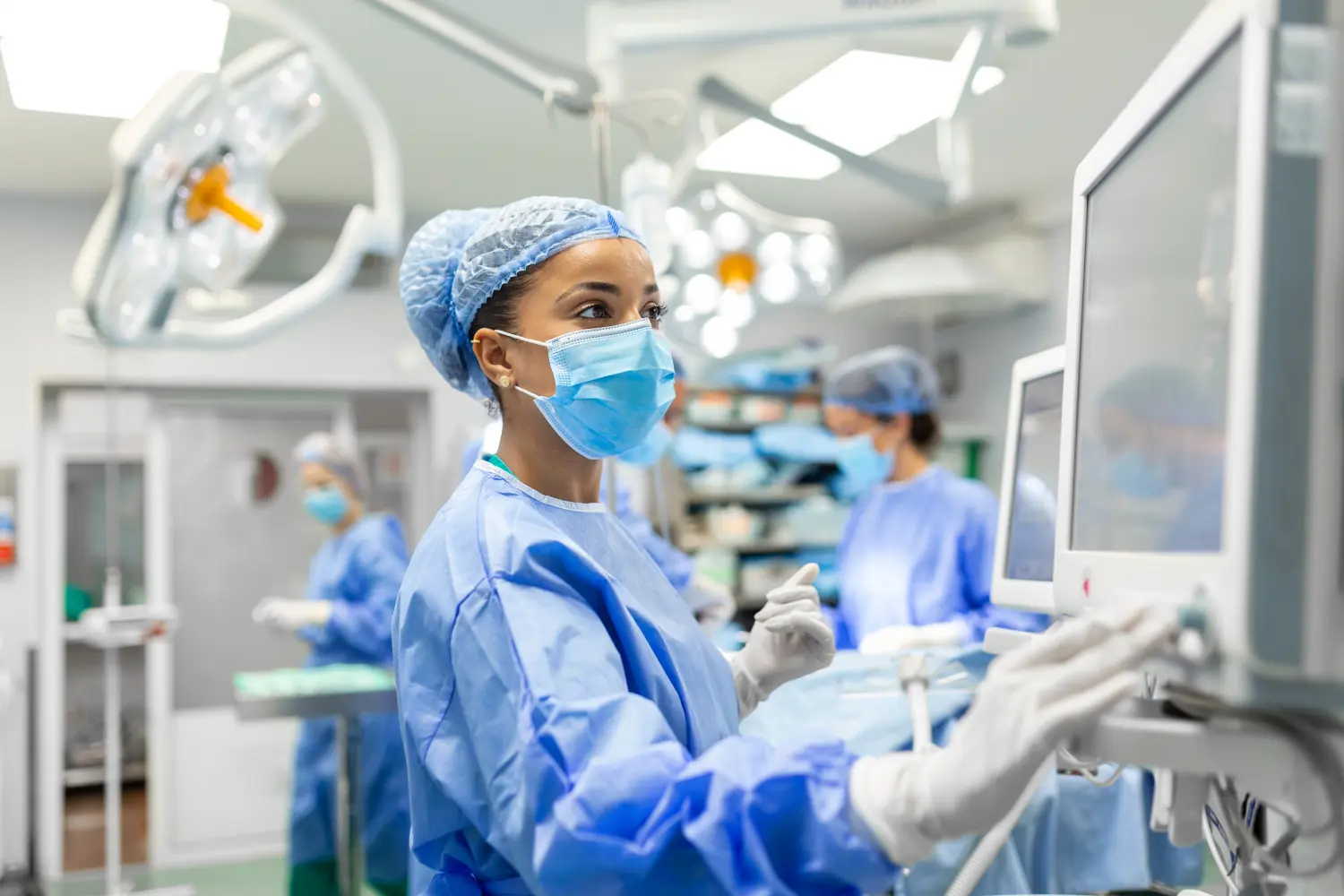 Anästhesist arbeitet im Operationssaal und trägt Schutzkleidung, während er Monitore überprüft, während er einen Patienten vor einem chirurgischen Eingriff sediert